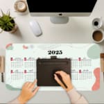 Infidu Calendar Design Desk Pad displaying a 2025 calendar layout with a light background and abstract red and gray accents. A writing pad on top of the desk pad