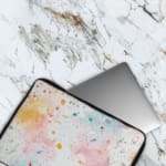 Colorful splatter paint design laptop sleeve with blue, orange, and pink splashes on a light background. The laptop sleeve covers the laptop