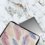 Infidu Pastel Feather Laptop Sleeve with soft pastel feathers in pink, purple, and blue on a light background, shown with office items. The laptop sleeve covers the laptop