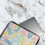 A colorful abstract laptop sleeve featuring pastel pink, blue, yellow, and green curved shapes on a white background with a zip closure. laptop sleeve kept on a plain white background The laptop sleeve covers the laptop