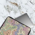 Laptop sleeve with topographic map design in pastel colors, featuring contour lines on a neutral background. The laptop sleeve covers the laptop