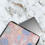 An abstract pastel laptop sleeve in pink, blue, and light green with circular and swirl patterns on a white background with a zip closure. The laptop sleeve covers the laptop