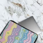 Retro-inspired laptop sleeve with wavy pastel stripes in pink, purple, yellow, green, and blue, covering the surface with a zip closure. The laptop sleeve covers the laptop