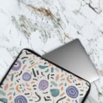 Organic abstract laptop sleeve with hand-drawn patterns in coral, purple, mint green, and black on a white background with a zip closure. The laptop sleeve covers the laptop