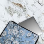 Elegant laptop sleeve with a blue background and white flowers with gray details, featuring small blossoms and leaves, secured with a zip. The laptop sleeve covers the laptop