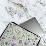 Laptop sleeve with purple floral design, featuring purple flowers and green leaves on a light background, with a secure zip closure. The laptop sleeve covers the laptop