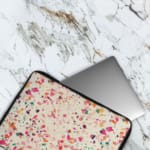 Laptop sleeve with a colorful terrazzo pattern in pink, orange, yellow, green, and black shapes on a white background, with a zip closure. The laptop sleeve covers the laptop