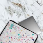 Laptop sleeve featuring a pastel terrazzo design with irregular shapes in pink, blue, turquoise, and dark shades on a light background. The laptop sleeve covers the laptop