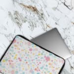 Laptop sleeve with colorful polka dots in pastel shades of pink, blue, yellow, and purple on a light background, with a secure zip closure. The laptop sleeve covers the laptop