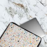 Laptop sleeve with a colorful speckled design in black, orange, blue, and yellow on a light background, featuring a secure zip closure. The laptop sleeve covers the laptop