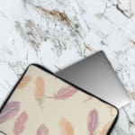 Infidu Pastel Feather Laptop Sleeve with a soft feather design in pastel shades on a light background, displayed on a white desk. The laptop sleeve covers the laptop