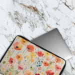 Colorful floral laptop sleeve with red, pink, yellow, and orange flowers on a light background. The laptop sleeve covers the laptop