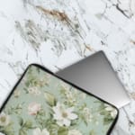 Floral laptop sleeve with white flowers and green leaves on a soft green background. The laptop sleeve covers the laptop