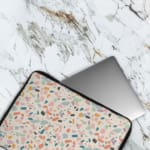 Terrazzo design laptop sleeve with blue, brown, and beige shapes on a light background, placed on a white desk with office supplies. The laptop sleeve covers the laptop